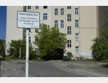 Mehrstöckiges, beiges Gebäude mit mehreren Fenstern, umgeben von Bäumen und Sträuchern. Im Vordergrund ist ein Parkplatz mit einem Schild, das das Abstellen von Fahrzeugen auf dem Privatgrundstück untersagt.