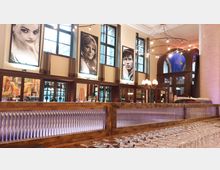 Großzügige Bar in einem stilvollen Interieur mit hohen Fenstern, dekorativen Lampen und großen schwarz-weißen Porträtbildern an den Wänden. Auf der Theke stehen zahlreiche fein ausgerichtete Weingläser, und im Hintergrund sind Holzverkleidungen und gläserne Vitrinen sichtbar.