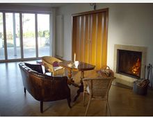 Ein Wohnzimmer mit hellem Holzboden, einem Kamin an der rechten Wand und einer Sitzecke mit einem Ledersofa und Korbstühlen um einen Holztisch. Im Hintergrund befinden sich große Glastüren, die auf eine Terrasse mit Ausblick ins Freie führen.