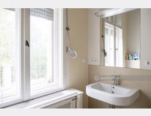 Helles Badezimmer mit einem großen Fenster, das zu einem Balkon mit einem dekorativen Geländer hinausführt. Ein Waschbecken mit Spiegel sowie eine Heizkörperabdeckung unter dem Fenster sind sichtbar.