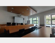 Ein moderner Besprechungsraum mit Holzinterieur, ausgestattet mit einem Konferenztisch, schwarzen Bürostühlen, einer weißen Projektionsfläche an der Wand und großen Fenstern, die viel Tageslicht hereinlassen. Im Hintergrund sind ein Flipchart und ein Projektor zu sehen.