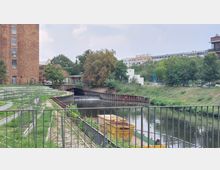 Ein städtischer Wasserkanal mit einer kleinen Stufe, umgeben von Grünflächen und modernen Gebäuden im Hintergrund. Links ist ein großes Backsteingebäude und rechts im Vordergrund ein Geländer sowie Baucontainer am Ufer.