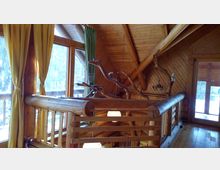 Innenraum einer Blockhütte mit sichtbarer Holzkonstruktion, großen Fenstern mit grünen und gelben Vorhängen, durch die Tageslicht und eine schneebedeckte Landschaft zu sehen sind. Im Vordergrund befinden sich ein hölzernes Geländer und verzweigte Äste als Gestaltungselemente.