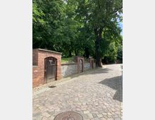 Eine gepflasterte Straße mit einer Steinmauer und mehreren roten Backsteintoren, die teilweise Holztüren enthalten. Im Hintergrund stehen große, dicht bewachsene Bäume, die die Umgebung beschatten.
