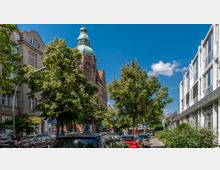 Straßenszene in einer Stadt mit einem historischen Gebäude im Hintergrund, das durch eine grüne Kuppel und aufwändige Architektur mit Fensterrosette auffällt. Links und rechts der Straße befinden sich Wohn- und Geschäftsgebäude; davor sind geparkte Autos und Bäume.