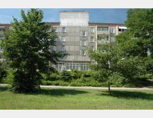 Wohnblock im Stil der Plattenbauweise mit mehreren Stockwerken und grauen Fassadenplatten, umgeben von einer Grünfläche mit Bäumen, Sträuchern und einem gepflasterten Weg im Vordergrund. Ein blauer Himmel bildet den Hintergrund.