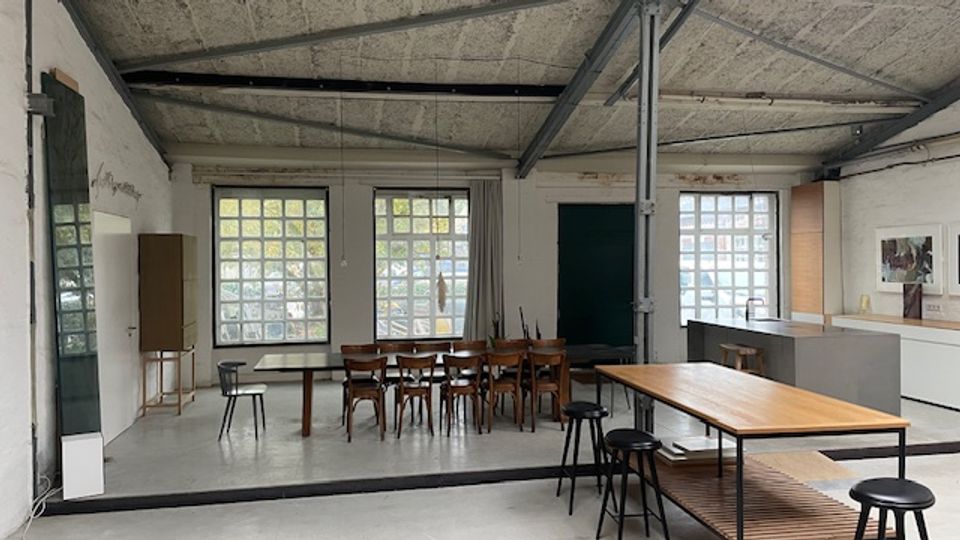 Großer, offener Raum im Industrie-Stil mit Sichtbetondecke, Stahlträgern und großen Glasbausteinfenstern, die viel Tageslicht hereinlassen. Im Inneren stehen mehrere Tische mit Stühlen und Hockern sowie eine lange Arbeitsfläche entlang der rechten Wand.