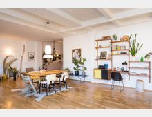 Helles Wohnzimmer mit Holzboden, einem großen Esstisch aus Holz mit sechs Stühlen und zwei Hängeleuchten darüber. An der rechten Wand befinden sich offene Holzregale mit Büchern, Pflanzen und Dekorationen, sowie ein gelb-schwarzes Sideboard; links verschiedene Pflanzen und Wandbilder.