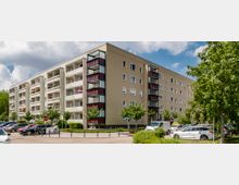 Mehrstöckiges Wohngebäude mit hellem Putz und mehreren Balkons, einige davon mit roten Verkleidungen und bepflanzt. Im Vordergrund geparkte Autos, kleine Bäume und ein Gehweg, im Hintergrund ein leicht bewölkter Himmel.