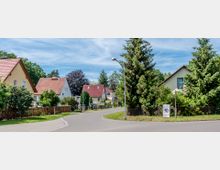 Wohngebiet mit einem asphaltierten Straßenverlauf, gesäumt von Einfamilienhäusern mit roten und braunen Dächern. Umgeben von grünen Bäumen, Gärten und Zäunen, unter einem klaren blauen Himmel.