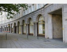 Straßenansicht eines Gebäudes mit heller Steinfassade und Arkaden, die auf Säulen ruhen. Unter den Bögen befinden sich große Fenster und Türen mit gelben Rahmen, und der Bürgersteig ist mit grauen Pflastersteinen ausgelegt.