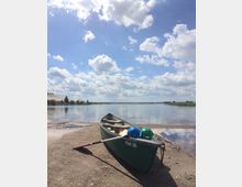 Ein grünes Kanu mit der Aufschrift "PAK 06" liegt auf einem sandigen Ufer an einem ruhigen See. Im Boot befinden sich zwei Kunststoffhelme, und ein Paddel lehnt an der Seite; im Hintergrund erstrecken sich flache Ufer und ein Himmel mit vielen Wolken.