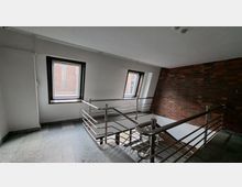 Innenliegendes Treppenhaus mit grauem Steinboden und Metallgeländer, daneben zwei rechteckige Fenster in einer schrägen Wand mit Blick auf eine Backsteinfassade. Eine Backsteinwand verläuft entlang des Treppenabgangs unter einer weißen Decke.