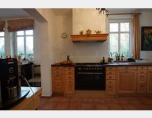 Küche mit hellen Holzschränken, einem schwarzen Herd in der Mitte und einer Dunstabzugshaube mit Dekoration. Rechts und links befinden sich große Fenster mit Blick auf einen Garten, darunter eine Fensterbank mit Flaschen und Dekorationsartikeln.