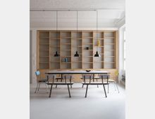 Minimalistisch eingerichteter Raum mit einer großen Holzregalwand im Hintergrund, einem langen Tisch mit mehreren Stühlen und drei schwarzen Hängelampen. Auf dem Tisch befinden sich vereinzelte Dekorationsobjekte, und das Tageslicht fällt durch ein großes Fenster von der rechten Seite ein.