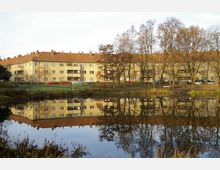 Ein mehrstöckiger Wohnblock mit gelber Fassade und roten Ziegeldächern, umgeben von kahlen Laubbäumen, spiegelt sich in einem ruhig daliegenden Teich im Vordergrund. Entlang der Straße vor dem Gebäude sind parkende Autos zu sehen, und die Umgebung wirkt grün und offen.