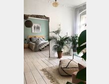 Helles Wohnzimmer mit Holzdielenboden und Teppich, dekoriert mit einer großen Zimmerpflanze und einem geometrischen Pouf. Im Hintergrund ein durchbrochener Türrahmen, der zu einem Schlafzimmer mit Bett und grüner Wand führt, verziert mit einem Schwarz-Weiß-Bild.