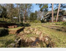 Garten mit unebener Landschaft, geprägt von Grasflächen, niedrigem Bewuchs und einer kleinen Teichmulde, umgeben von Backsteinmauern; im Hintergrund ein weißes Wohngebäude mit Schrägdach und ein Pavillon unter hohen Bäumen.