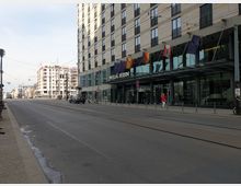 Straßenansicht eines modernen Hotelgebäudes mit der Aufschrift „Meliá Berlin“. Vor dem Gebäude hängen mehrere Fahnen, und Straßenbahnschienen führen entlang der breiten Straße, die von einer gehenden Person und parkenden Fahrrädern gesäumt ist.