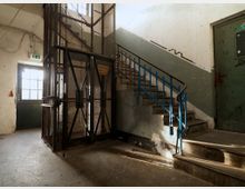 Industrieartiges Treppenhaus mit abgenutzten Details, bestehend aus einer Metalltreppe mit blauem Geländer, einem Drahtkäfig-Aufzug und einer grünen Tür. Die Umgebung wirkt alt und ist teilweise von Graffiti und Abnutzungsspuren geprägt.