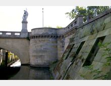 Steinbrücke mit einem Rundturm und einer Statue auf einem Sockel, flankiert von einer begrünten Uferbefestigung aus gealtertem Mauerwerk; darunter fließt ein ruhiges Gewässer.