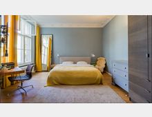 Schlafzimmer mit einem Doppelbett mit gelber Decke vor einer blau gestrichenen Wand, flankiert von zwei Wandleuchten. Links stehen ein Schreibtisch mit einem Stuhl und geöffneten Fenstern mit gelben Vorhängen; rechts befinden sich eine graue Kommode und ein Schrank.