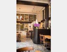 Ein gemütliches Café mit Holztischen und -stühlen, einer Theke mit Blumen in Vasen und Regalen im Hintergrund, die mit Flaschen und Dekorationsgegenständen gefüllt sind. Die helle Decke wird durch hängende Lampen akzentuiert, und der Boden ist mit gemusterten Fliesen gestaltet.