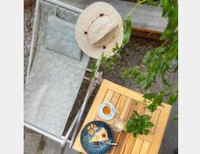 Ein kleiner Außenbereich mit einem Holzklapptisch, darauf ein Teller mit einem Kuchenstück, eine Tasse Tee, ein Glas Wasser und eine kleine Topfpflanze. Daneben steht eine Liege mit floralem Muster, auf der ein Strohhut liegt; Pflastersteine und grüne Äste ergänzen die Umgebung.