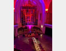 Innenraum einer modern umgestalteten Kirche mit hoher Decke, beleuchtet in violetten und roten Tönen. Im Vordergrund steht eine lange, gedeckte Tafel, während der Altarbereich von einer großen, kunstvoll gestalteten Installation aus Linien und Glas geprägt ist.