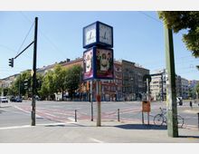 Straßenkreuzung mit Fußgängerüberweg und Ampelanlagen in einem städtischen Umfeld. Im Vordergrund steht eine Uhr mit Werbeanzeigen, im Hintergrund sind mehrstöckige Wohn- und Geschäftsgebäude sowie vereinzelt Bäume sichtbar.