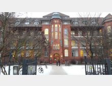 Backsteinfassade eines mehrstöckigen Gebäudes mit symmetrischer Gestaltung und Rundbogenfenstern, beleuchteten Innenräumen und Mansardendach. Im Vordergrund ein schneebedeckter Hof mit Fahrrädern und zwei Personen vor einem Eingang mit Tor.