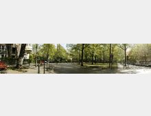 Eine grüne Parkanlage in einer städtischen Umgebung, umgeben von Wohngebäuden. Gepflasterte Wege, rote Parkbänke und zahlreiche Bäume prägen die Szenerie, während im Hintergrund geparkte Autos und Straßenschilder sichtbar sind.