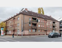 Wohnhaus aus der Nachkriegszeit mit braun-beiger Fassade und roten Klinkerelementen an einer Straßenecke. Im Vordergrund eine Kreuzung mit Ampeln, geparkten Fahrzeugen und Fußgängern; herbstliche Bäume und Werbesäule am Gehweg.