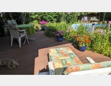 Holzterrasse in einem Garten mit einem Liegestuhl, einer Sitzgruppe mit grünem Tischtuch und mehreren blühenden Blumenkübeln; im Hintergrund ein Swimmingpool und dichte grüne Vegetation. Ein kleiner Hund liegt im Schatten auf der Terrasse.