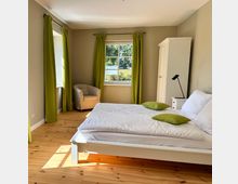 Helles Schlafzimmer mit Holzboden, einem weißen Bett mit grünen Dekokissen, einem weißen Kleiderschrank und einem Sessel in der Ecke. Die großen Fenster haben grüne Vorhänge, durch die Tageslicht hereinfällt, mit Blick auf eine grüne Außenlandschaft.