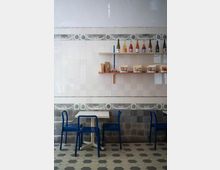 Ein kleiner Innenraum mit hellen Wandfliesen, die oben weiß und unten grau gestaltet sind, verziert mit einem ornamentalem Muster. Vor der Wand stehen ein niedriger Tisch mit blauer Bestuhlung sowie ein Regal mit Flaschen und Lebensmitteln, darunter helle Verpackungen. Der Boden ist mit sechseckigen Fliesen in Schwarz, Weiß und Grau gestaltet.