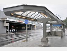 Eingangsbereich der U-Bahn-Station "Rathaus Spandau" mit überdachter Treppenanlage aus Glas und Metall. Im Hintergrund eine Überführung sowie eine nasse Straße mit Fußgängern und Fahrradschild.