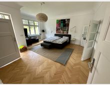 Ein helles Schlafzimmer mit Fischgräten-Holzfußboden, einem Doppelbett mit dunklem Gestell, einer modernen Hängelampe und einem großen Fenster, das viel Tageslicht hereinlässt. An der Wand hängt ein buntes Gemälde, und daneben stehen Nachttische mit Leselampen.