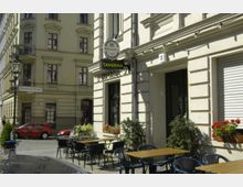 Straßenszene mit einem Gebäude im klassizistischen Stil, das ein Restaurant mit dem Schild "Taverna" beherbergt. Davor stehen leere Holztische mit schwarzen Stühlen auf dem Gehweg, umgeben von Blumenkästen und Pflanzen; im Hintergrund eine ruhige Kreuzung mit einem Straßenschild und geparkten Autos.