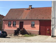 Ein einstöckiges Gebäude mit hellrosa Fassade und rotem Ziegeldach, links einige Fenster und eine Tür mit kleinen Treppenstufen davor, rechts ein großes rotes Holztor mit der Aufschrift „Luisenhof im Fischersteig“. Davor führt eine gepflasterte Straße entlang.