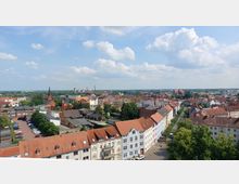 Luftaufnahme einer Kleinstadt mit rotgedeckten Dächern, historischen Gebäuden und einer Kirche mit spitzem Turm in der Mitte. Im Hintergrund sind Industriebauten und flache Landschaften unter einem Himmel mit lockerer Bewölkung zu sehen.