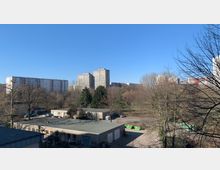 Blick auf eine städtische Landschaft mit mehreren Hochhäusern im Hintergrund, umgeben von kahlen Bäumen und Grünflächen. Im Vordergrund befinden sich einstöckige Gebäude mit Flachdächern und einige grüne Müllcontainer.