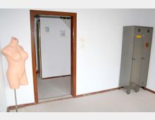 Ein schlichter Raum mit weiß gestrichenen Wänden, einem Türrahmen aus dunklem Holz, der in einen angrenzenden Bereich mit heller Teppichauslage führt. Links steht eine nackte Schaufensterpuppe auf einem Ständer, rechts befindet sich ein grauer Metallschrank mit Schließfächern.