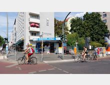 Straßenszene an der Kreuzung Grünberger Straße in einer Stadt, mit einem Eckgebäude, auf dessen Fassade rosa Sonnenschirme und ein Wandrelief zu sehen sind. Im Erdgeschoss befindet sich ein kleiner Kiosk mit der Aufschrift "Eastside Kiosk", daneben fahren mehrere Radfahrer und im Hintergrund sind Bäume sowie weitere Gebäude zu erkennen.