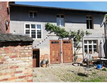 Ein kleiner Innenhof mit Pflastersteinen vor einem zweistöckigen Gebäude mit grauer Fassade und Holztüren. An der Wand ranken Pflanzen, und links im Vordergrund ist ein Gebäude aus rotbraunem Backstein zu sehen; im Hof stehen Gartenmöbel und ein Grill.