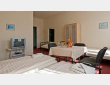 Ein einfach eingerichtetes Mehrbettzimmer mit mehreren Einzelbetten, einem Tisch mit vier Stühlen, einem kleinen Fernseher auf einem Regal und einem Kleiderschrank. Die Wände sind hell, der Boden rot, und das Zimmer wird von schlichter Dekoration wie Bildern und einer Blumenvase ergänzt.