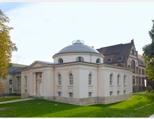 Heller, klassizistischer Bau mit einer Kuppel, hohen Rundbogenfenstern und einem Eingang mit Säulenportal, umgeben von einer Grünfläche. Im Hintergrund stehen weitere historische Gebäude mit Ziegelfassaden.