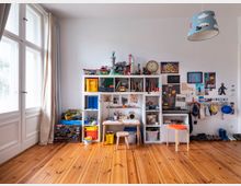 Ein Kinderzimmer mit einem großen weißen Regal an der Wand, gefüllt mit Spielzeug, Büchern und Bastelutensilien. Darunter ein kleiner Tisch mit Stühlen, daneben hängen Taschen und Hüte an einer Wandleiste; Holzdielenboden und ein großes Fenster mit weißen Vorhängen.