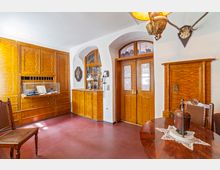 Historischer Raum mit hellen Holzmöbeln, darunter ein großer eingebauter Schrank mit Glasvitrine, Holztüren mit verzierten Glaseinsätzen und ein runder Holztisch mit Spitzendecke. An den Wänden sind dekorative Elemente wie ein Telefon und ein Hirschgeweih angebracht.