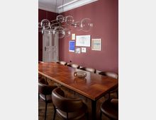 Esszimmer mit einer langen rechteckigen Holztischplatte in Parkettoptik, umgeben von gepolsterten braunen Stühlen. Die Wände sind dunkelrot gestrichen und mit gerahmten Kunstwerken geschmückt, eine große Pendelleuchte mit Glas-Kugeln hängt von der Decke.